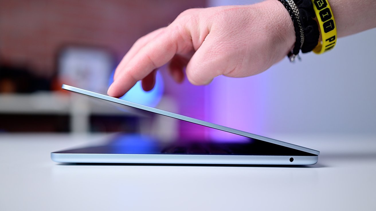 Hand touching partially closed laptop on white surface, wearing black and yellow wristbands, with blurred background and purple lighting.