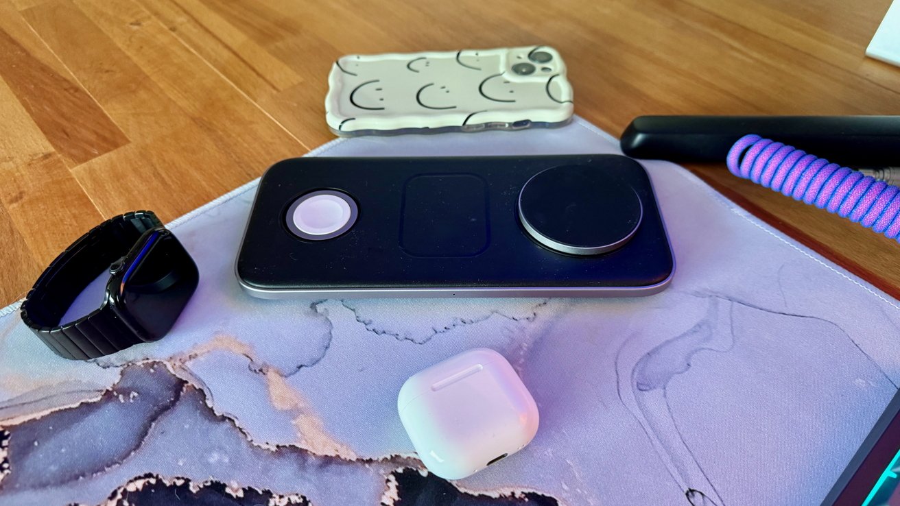 A black smartwatch, white phone case, wireless charger, AirPods, and coiled purple cable lie on a desk with a marble-patterned mouse pad.
